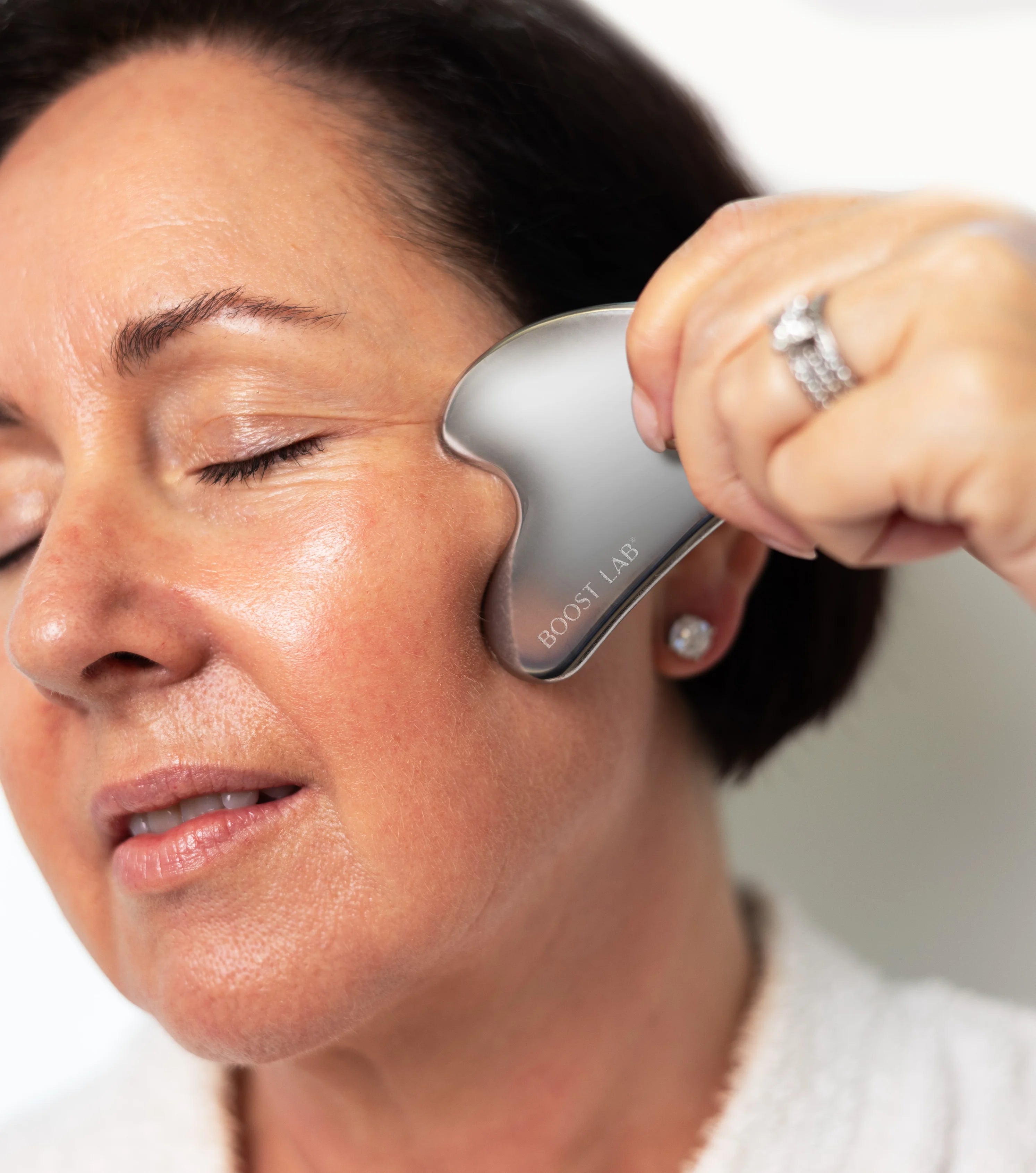 A close-up of a woman using a silver Boost Lab facial massage tool on her temple, with her hand holding the tool and a ring visible on her finger.